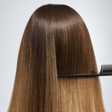 A woman with long brown hair is holding a black comb. The comb is going through her hair. The woman is facing away from the camera.