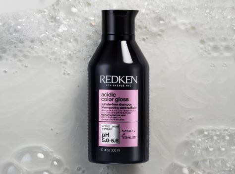 A bottle of Redken Acidic Color Gloss Sulfate-Free Shampoo sits in a bed of pink bubbles.