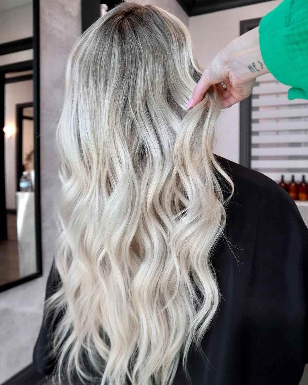  A client with long, wavy platinum blonde hair is shown from behind in a salon chair, with a stylist's hand examining a section of her hair