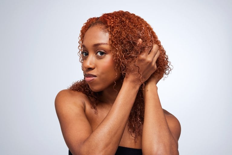 A woman with coily, vibrant auburn hair holds her curls, showcasing their definition and texture against a light gray backdrop. This image highlights healthy, well-maintained curls.