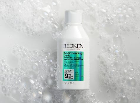 A bottle of Redken Acidic Bonding Concentrate Silicone-Free Shampoo sits in a bed of white bubbles.