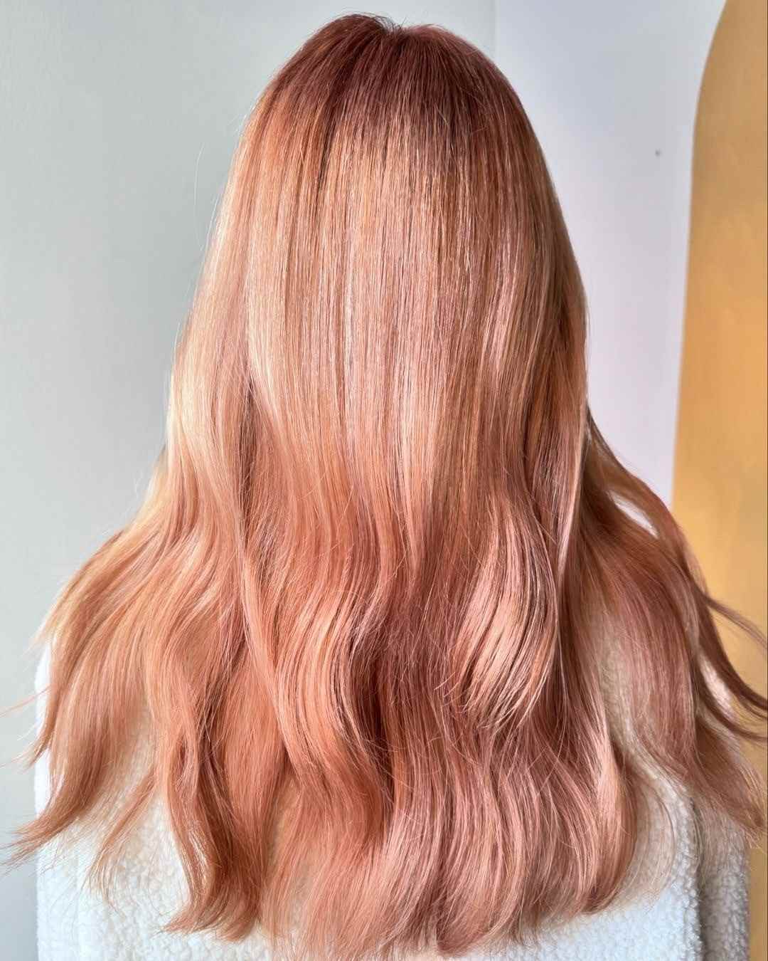 A person's long, wavy hair in a beautiful rose gold or peachy blonde color is displayed from behind